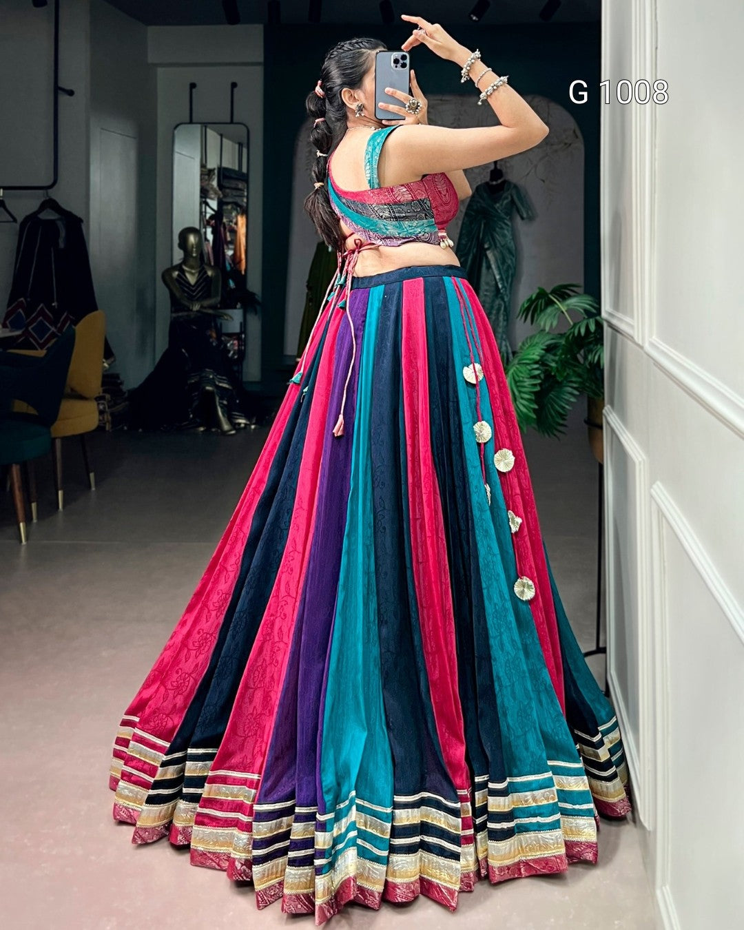 A woman takes a mirror selfie in a clothing boutique wearing a vibrant, multicolored traditional Indian lehenga choli set. The skirt features vertical stripes of magenta pink, teal blue, and purple, adorned with silver coin embellishments, and finished with a striped border.