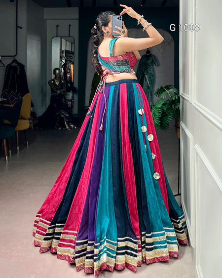 A woman takes a mirror selfie in a clothing boutique wearing a vibrant, multicolored traditional Indian lehenga choli set. The skirt features vertical stripes of magenta pink, teal blue, and purple, adorned with silver coin embellishments, and finished with a striped border.