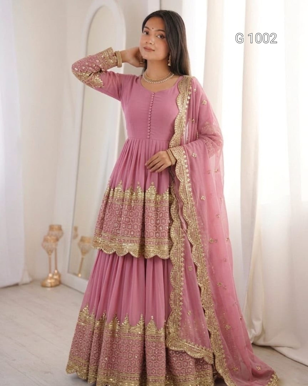 A woman models a pink traditional ethnic outfit with gold embroidery, featuring a long-sleeved kurta, flared skirt, and matching dupatta, set against a soft cream-colored indoor background.