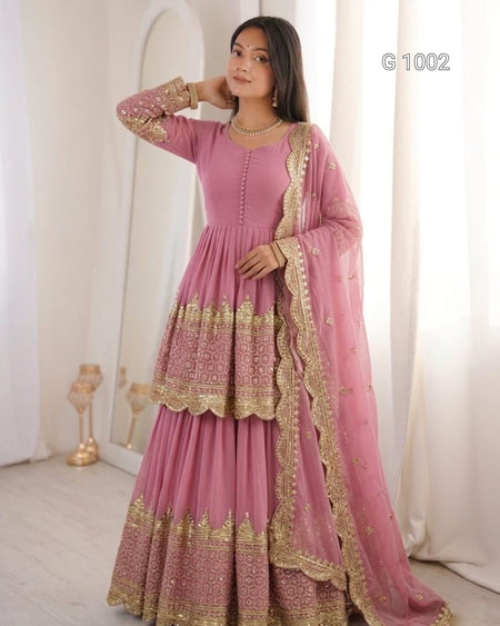 A woman models a pink traditional ethnic outfit with gold embroidery, featuring a long-sleeved kurta, flared skirt, and matching dupatta, set against a soft cream-colored indoor background.