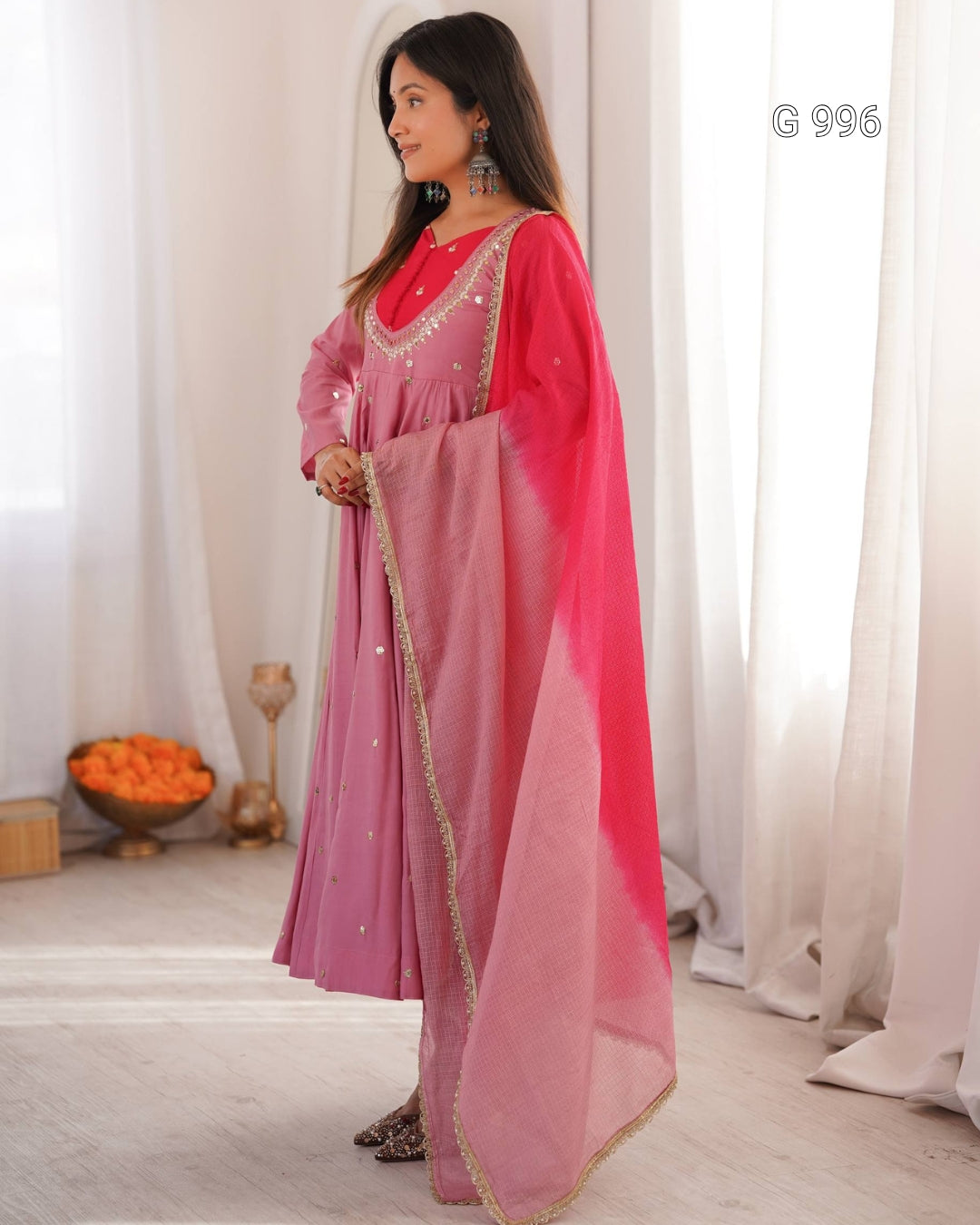 A woman models a traditional Indian outfit consisting of a pink kurta with gold embroidery, a matching pink skirt, and a bright pink dupatta draped over her arm, set against a background of white curtains.