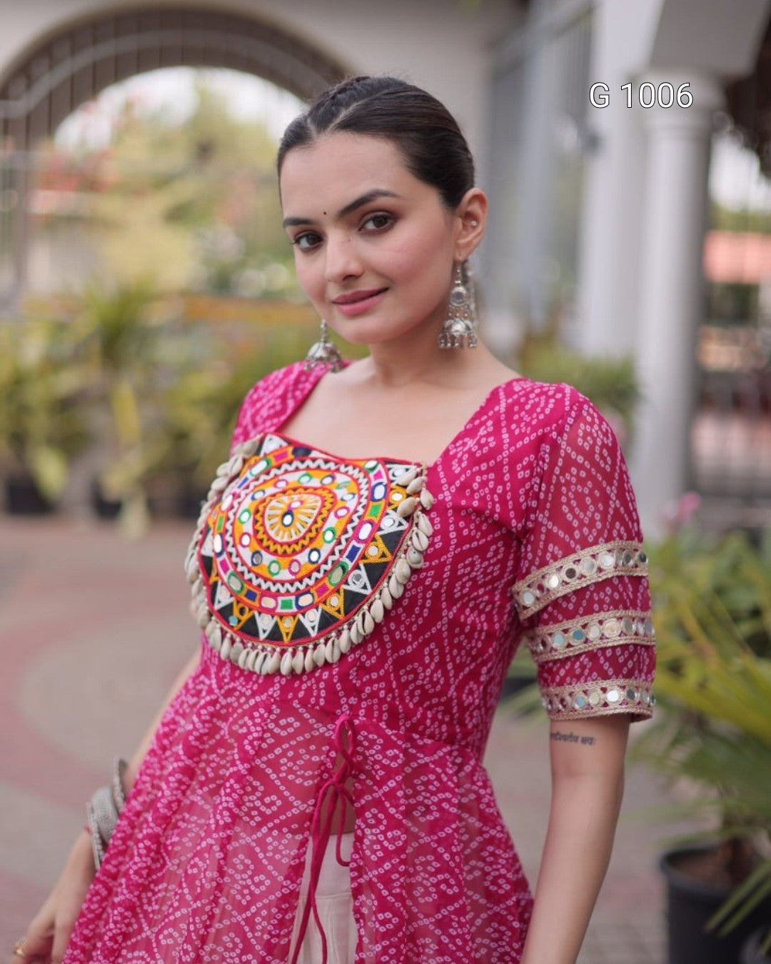 A woman wearing a bright pink printed kurta with a heavily embroidered multicolor yoke and mirrored sleeve bands, photographed outdoors in a softly blurred courtyard setting.
