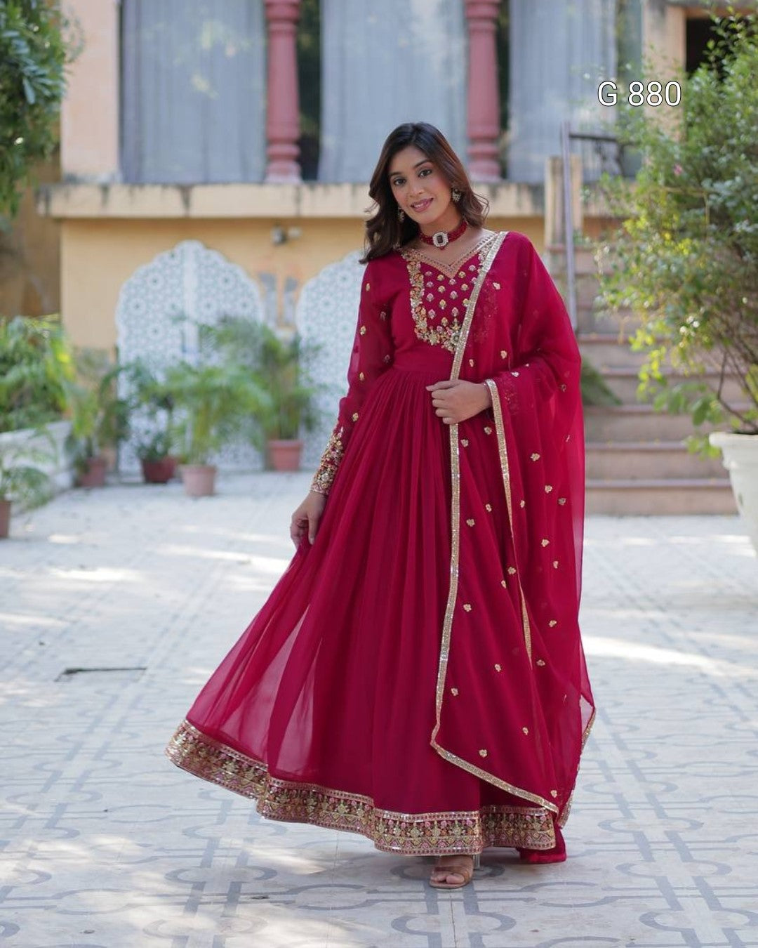 Woman in a red traditional outfit standing outdoors with greenery and a building in the background.