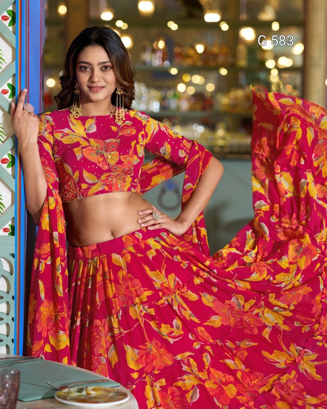 A woman models a bright pink and orange floral print lehenga choli set in an indoor setting with warm bokeh lights in the background. The set features a matching printed dupatta.
