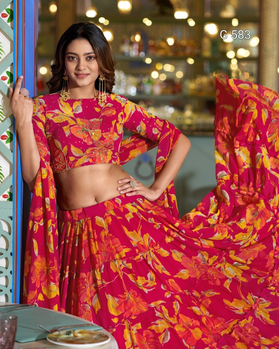 A woman models a bright pink and orange floral print lehenga choli set in an indoor setting with warm bokeh lights in the background. The set features a matching printed dupatta.