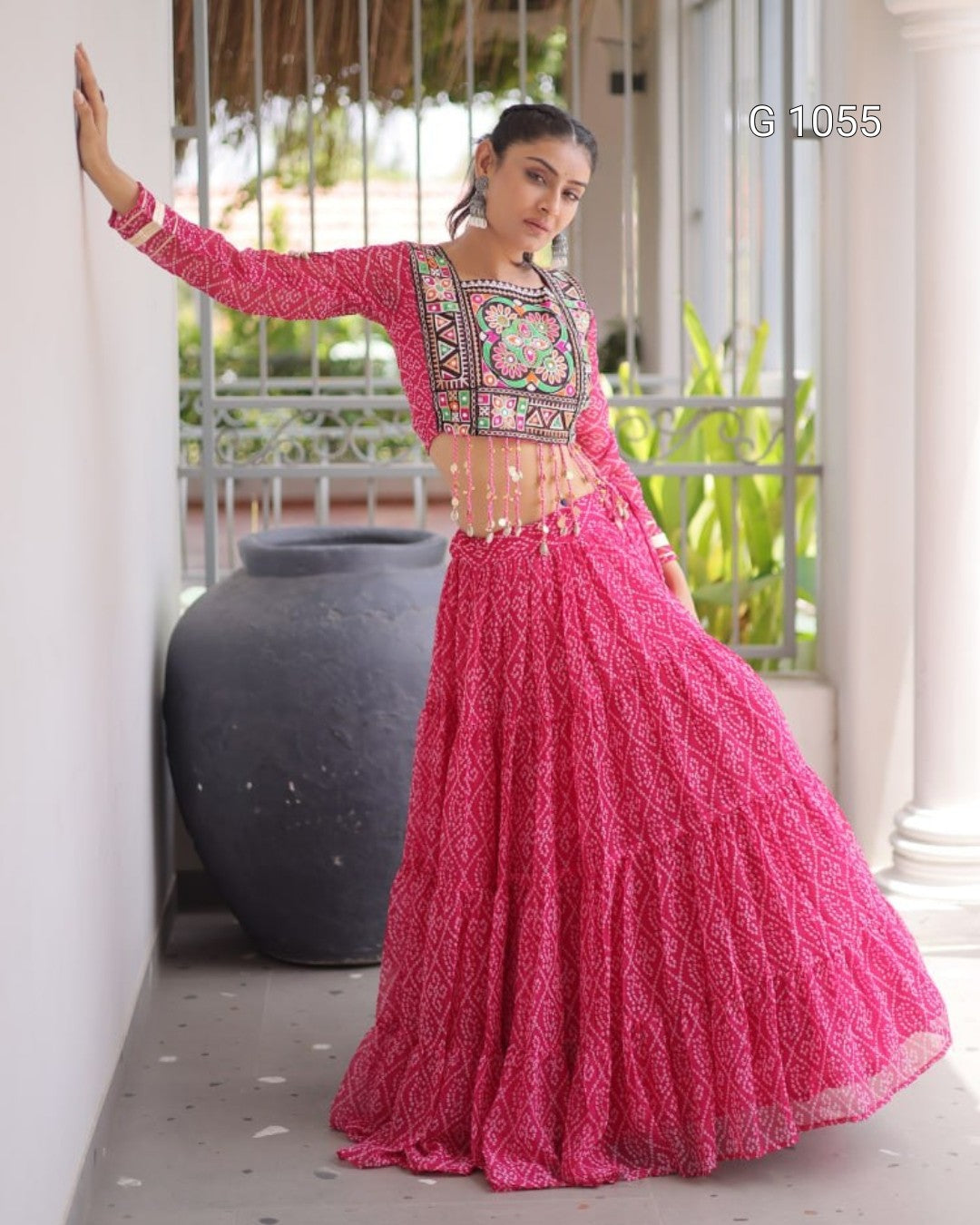 A woman modeling a bright pink traditional Indian outfit with a heavily embroidered neckline and a full flared skirt, standing on a porch with a large dark pot behind her.