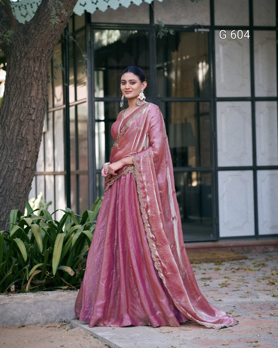 Woman in a pink saree standing outdoors with a building and plants in the background.