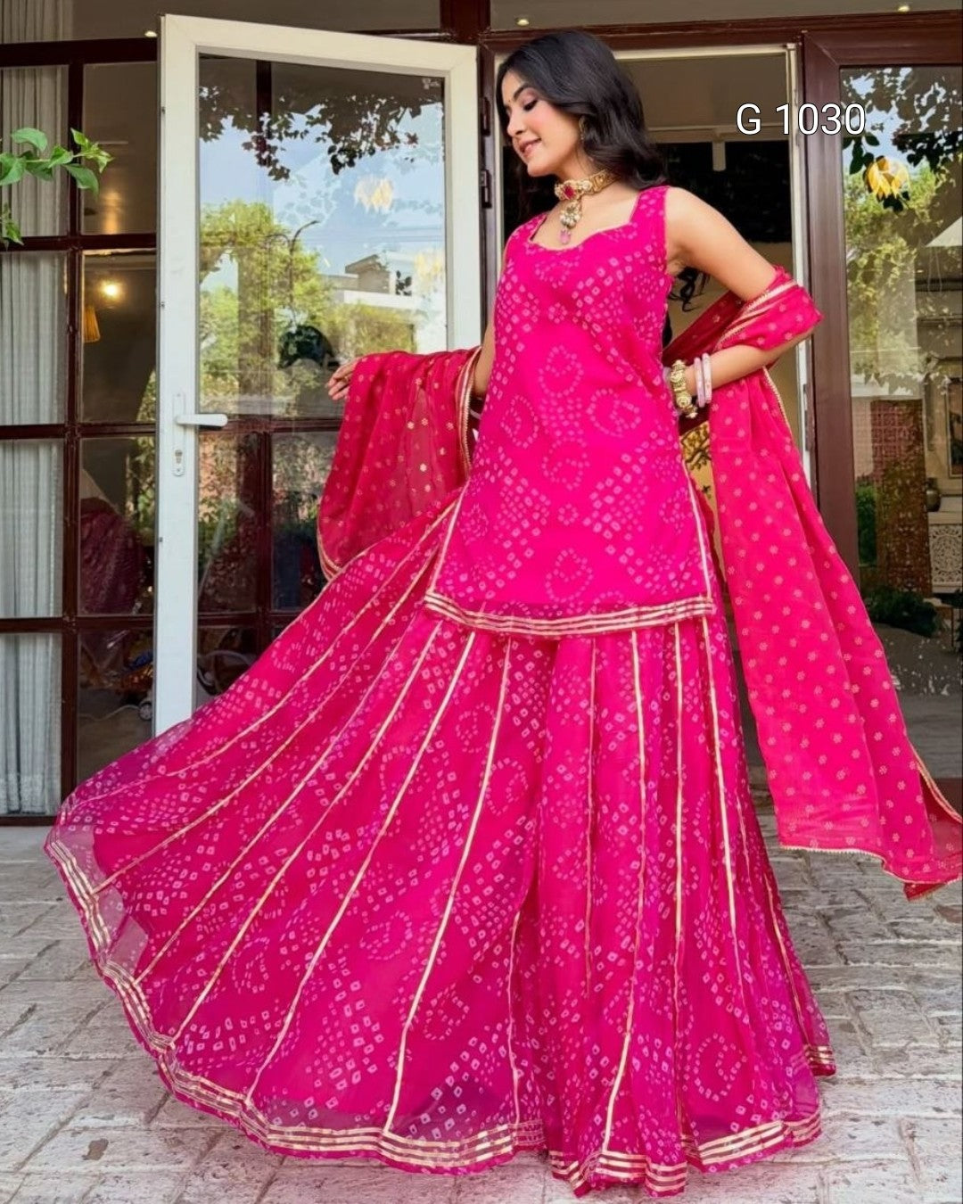 A woman modeling a bright pink traditional Indian outfit with matching dupatta, featuring gold border details and printed motifs, standing in front of a glass door.