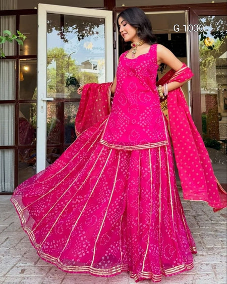 A woman modeling a bright pink traditional Indian outfit with matching dupatta, featuring gold border details and printed motifs, standing in front of a glass door.