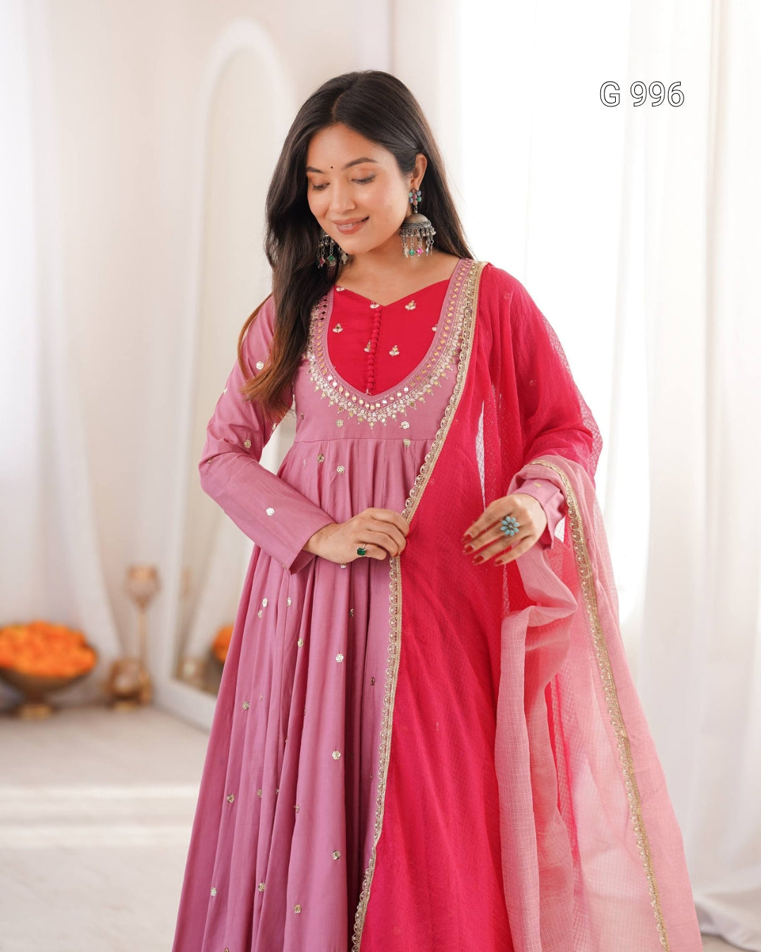 A woman modeling a traditional Indian outfit consisting of a pink long kurta with silver embroidery, a matching pink dupatta, and a bright pink outer layer draped over her shoulder, set against a soft white indoor background.