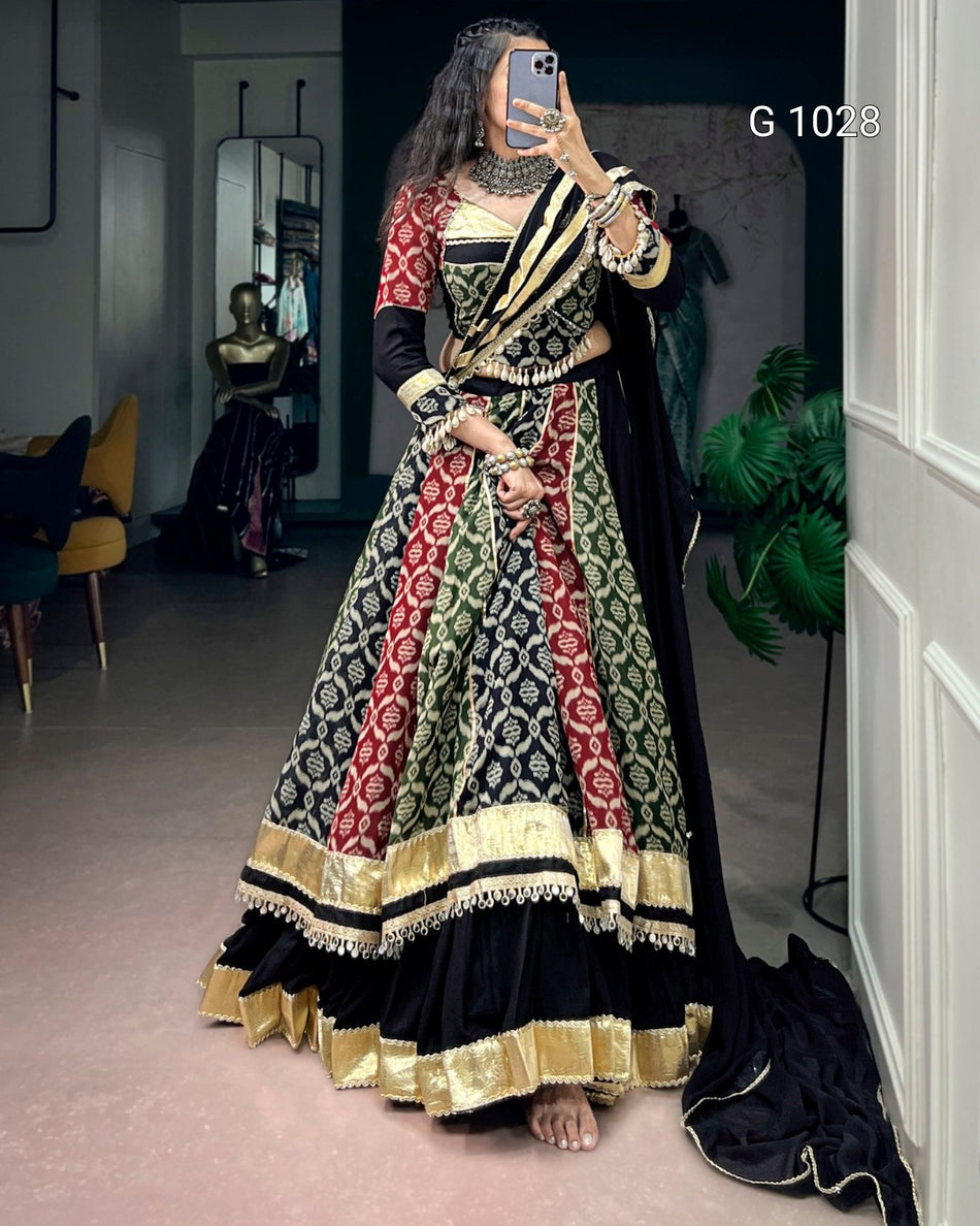 A woman takes a mirror selfie wearing a traditional Indian Anarkali gown with a black base, red and green patterned vertical panels, and gold zari embroidery. The dress features a flared skirt, a fitted bodice, and a draped dupatta.