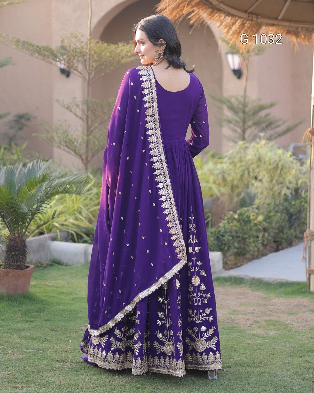 A woman wearing a purple traditional ethnic outfit with gold embroidery, standing outdoors in a garden setting. The outfit includes a long-sleeved kurta, flared pants, and a matching dupatta.