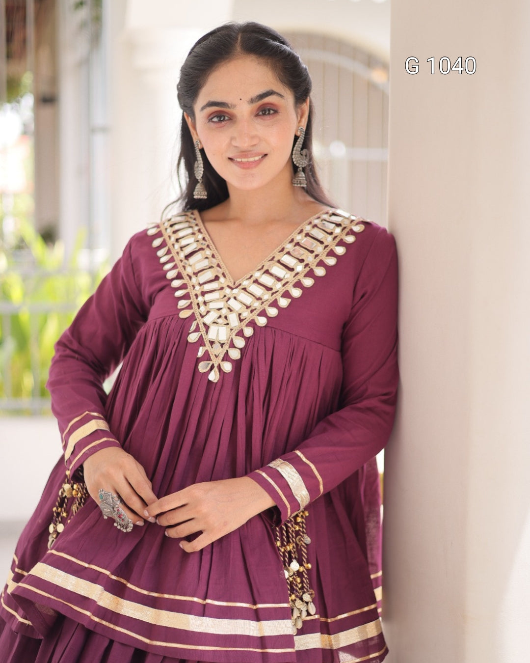 A woman wearing a deep maroon Anarkali-style kurta with gold and cream embroidery around the V-neck and cuffs, leaning against a light-colored wall in a softly blurred indoor setting.