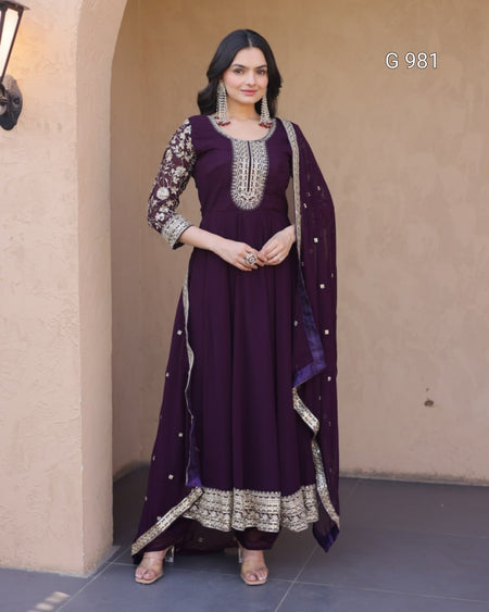 A woman models a deep purple traditional Indian outfit with silver embroidery, featuring a long kurta, matching trousers, and a dupatta, set against a beige stucco wall.