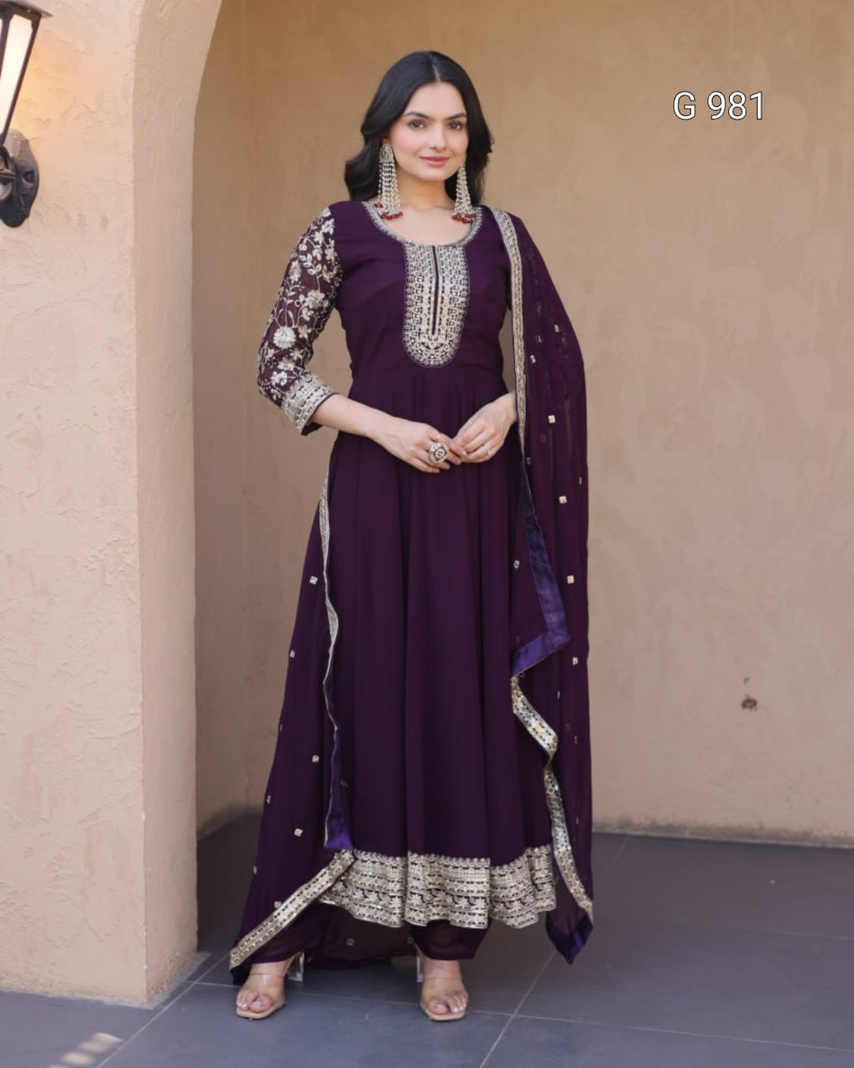 A woman models a deep purple traditional Indian outfit with silver embroidery, featuring a long kurta, matching trousers, and a dupatta, set against a beige stucco wall.