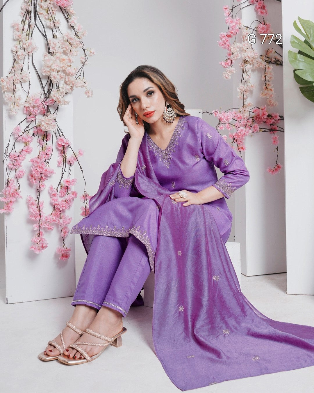 Woman in a purple traditional outfit sitting against a white background with pink floral decorations.