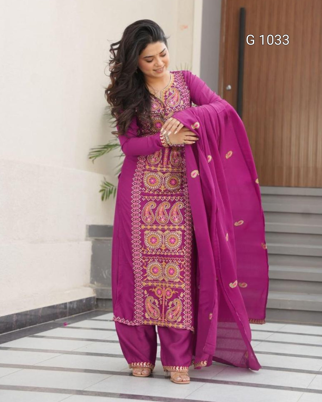 A woman modeling a traditional Indian salwar kameez suit in magenta purple with intricate gold and green embroidery, paired with matching pants and a sheer dupatta, standing in front of a modern building wall.