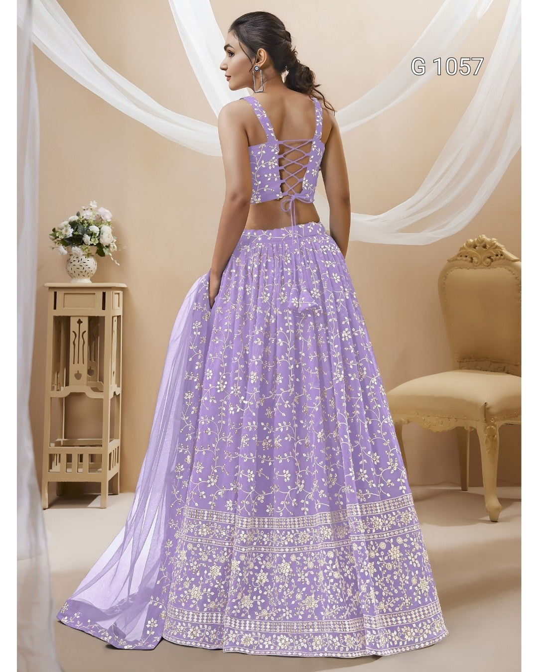 A woman modeling a lavender purple embroidered lehenga choli set with a matching dupatta, shown from the back in a decorated indoor studio setting.