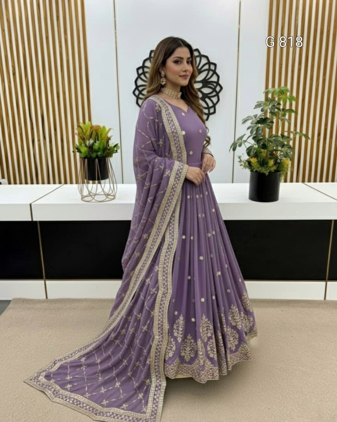 Woman in a purple saree with gold details standing in a room with plants and a decorative wall.