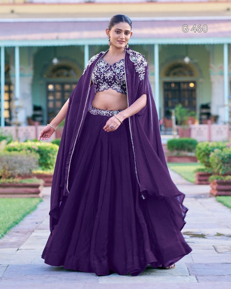 A woman models a deep purple traditional Indian outfit consisting of a flared skirt, an embroidered crop top, and a matching draped shawl, set against a background of a historic building and green garden.
