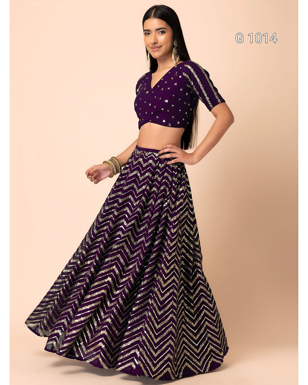 A woman modeling a traditional Indian outfit consisting of a dark purple cropped blouse with silver embellishments and a matching flared skirt with a purple, black, and silver chevron pattern, set against a solid beige background.