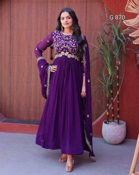 Woman in a purple traditional outfit standing against a red wall with a plant.