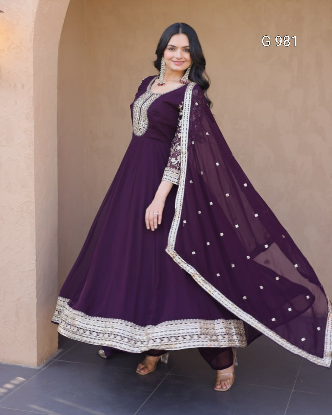 A woman modeling a deep purple traditional Anarkali gown with intricate silver embroidery and a matching sheer dupatta draped over her arm, set against a beige stone archway background.
