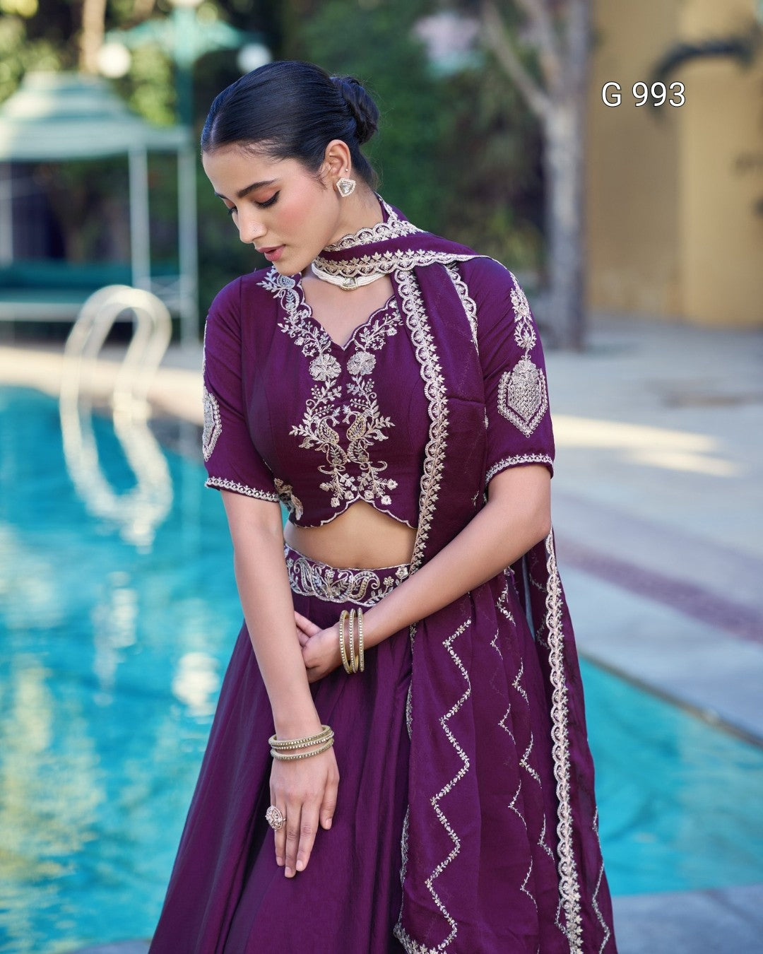 A woman wearing a deep purple traditional outfit with intricate gold embroidery, standing beside a swimming pool. The outfit features a short-sleeved crop top with a keyhole neckline and a matching flared skirt.