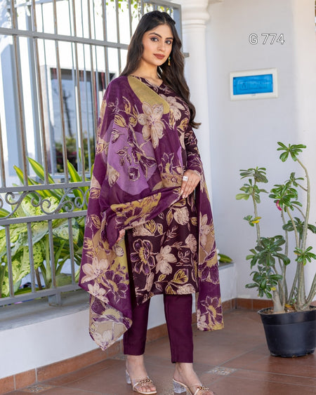 Woman wearing a purple floral outfit standing indoors with plants and a window in the background