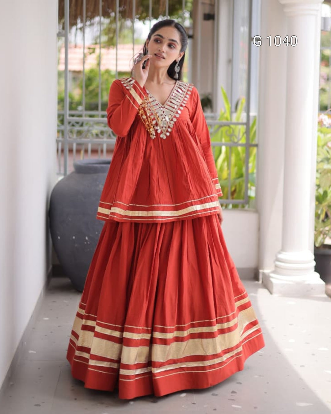 A woman models a traditional red Indian outfit with beige and gold striped borders, standing on a covered walkway with white pillars and green plants in the background.