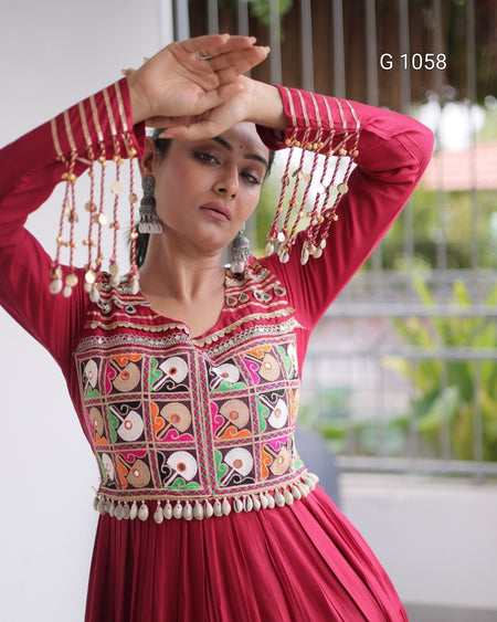 A woman wearing a deep red Anarkali-style dress with intricate multicolor embroidery and gold-toned hanging tassels on the sleeves, posing with her hands above her head. The background shows a softly blurred indoor/outdoor setting with a white wall and greenery.