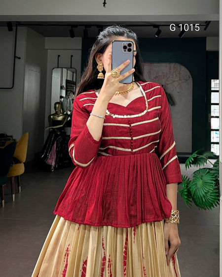 A woman takes a mirror selfie wearing a red and beige striped traditional-style top with a pleated waist, paired with a beige pleated skirt featuring red tie-dye patterns. She is wearing gold jewelry and holding a gray smartphone.