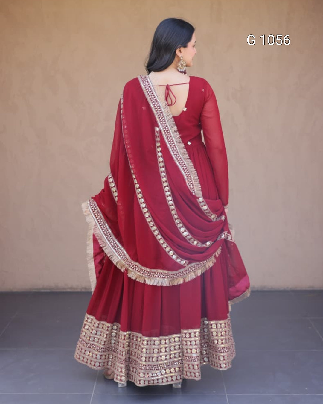 A woman wearing a deep red traditional ethnic outfit with gold embroidered borders, shown from the back against a plain beige wall.