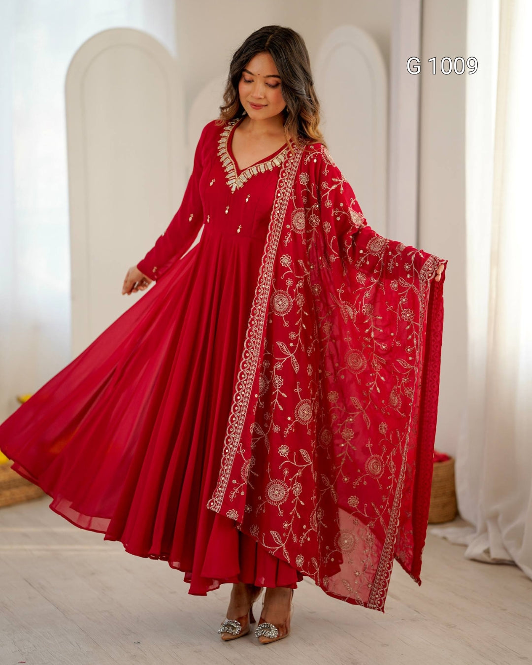 A woman modeling a traditional red Indian outfit with a long flared skirt, embellished neckline, and a matching embroidered dupatta draped over one arm in an indoor setting with white arches and curtains.
