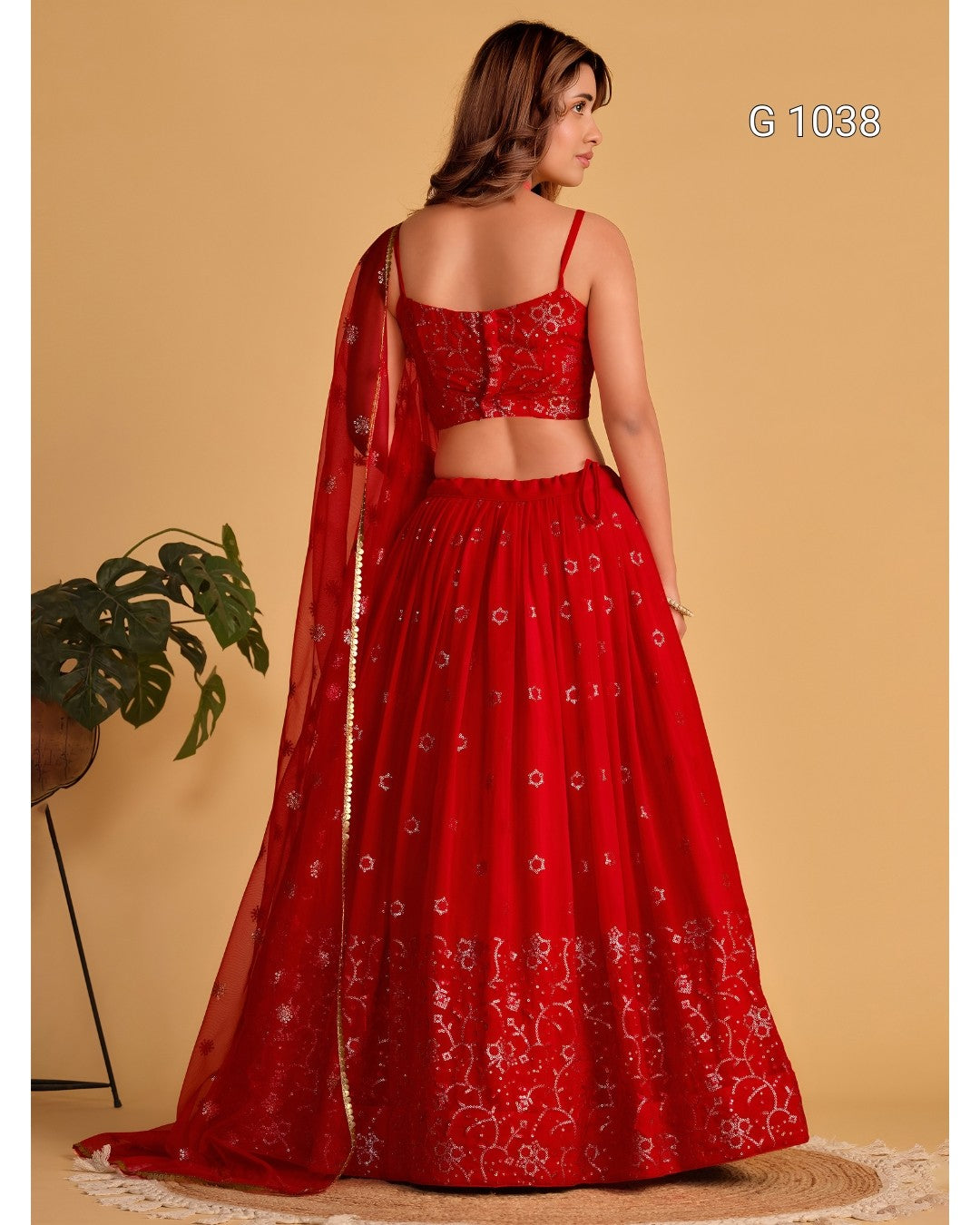 A woman modeling a red embroidered Indian ethnic outfit with a flared skirt, fitted crop top, and matching sheer dupatta, shown from the back against a warm beige studio background.