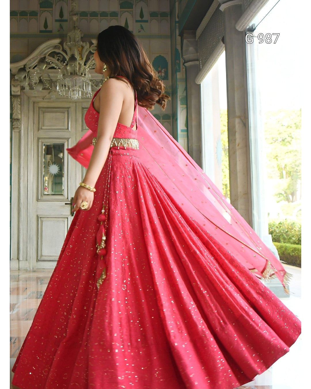 A woman wearing a bright pink lehenga choli with gold embellishments and a matching dupatta, photographed indoors near large windows.