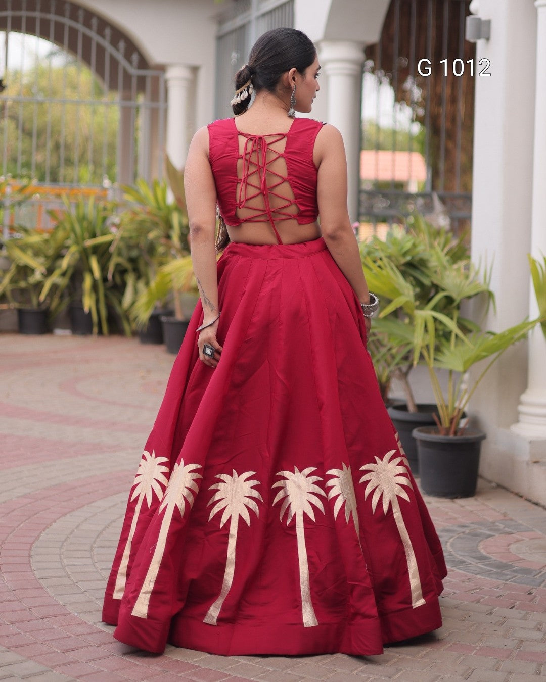 A woman wearing a deep red traditional Indian dress with a high-waisted flared skirt and lace-up back, photographed from behind in an outdoor courtyard setting.