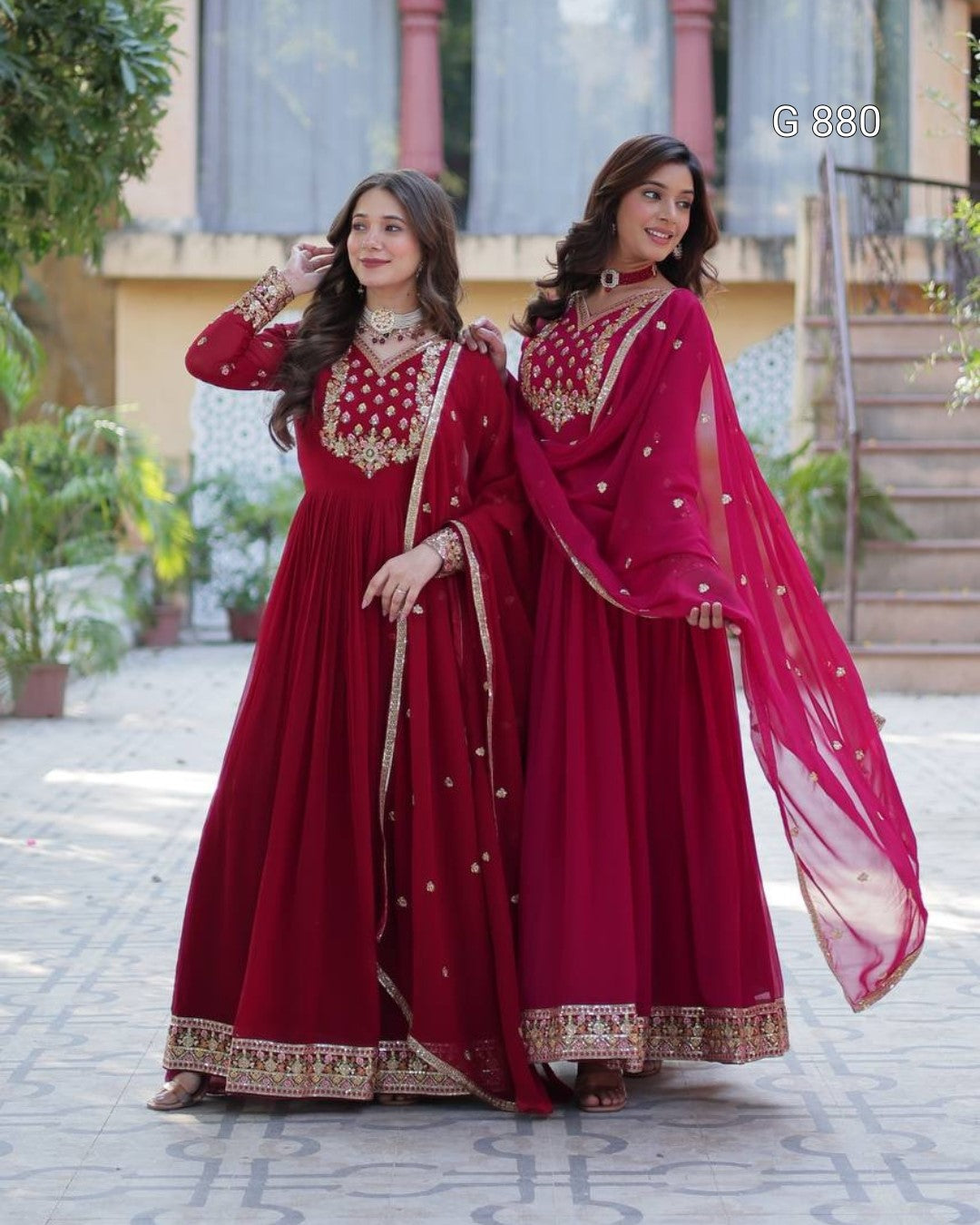 Two women in red traditional outfits standing outdoors with a building and plants in the background.