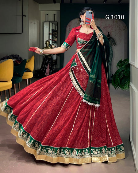 A woman modeling a traditional Indian red lehenga choli with a green and black dupatta, featuring intricate gold and green border patterns, in a mirror selfie.