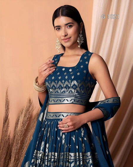 A woman modeling a dark teal blue and silver embellished Indian ethnic outfit, featuring a sleeveless crop top, flared skirt, and dupatta, set against a warm beige backdrop with draped curtains.