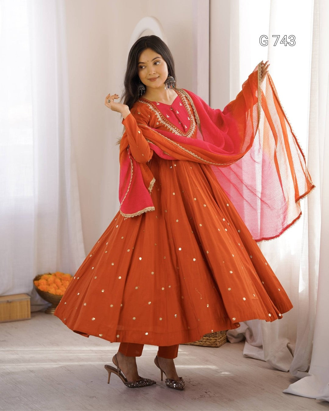 Woman wearing an orange and pink traditional outfit with a sheer dupatta in a well-lit room.