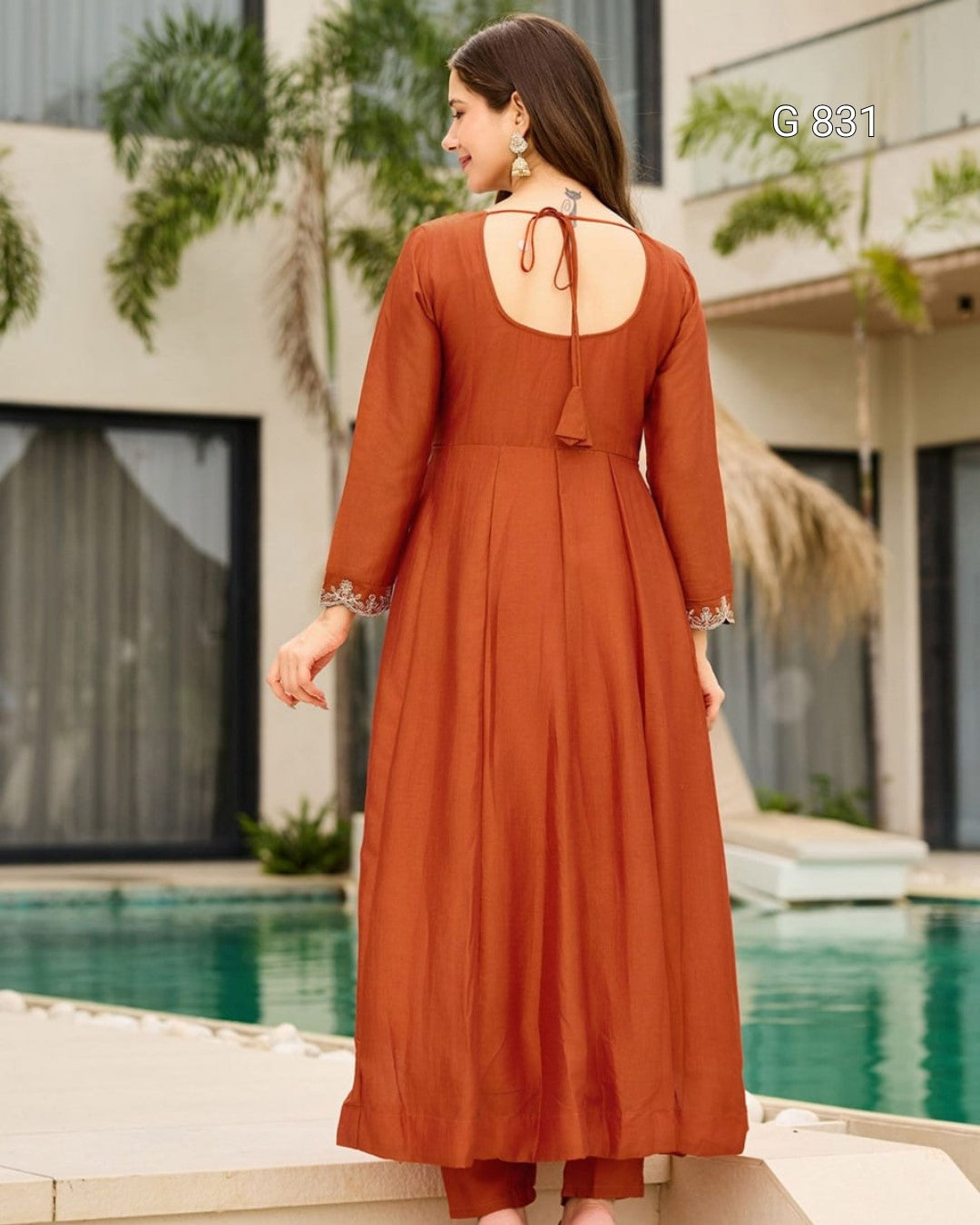 Woman in a rust-colored dress standing by a pool with a building and plants in the background.