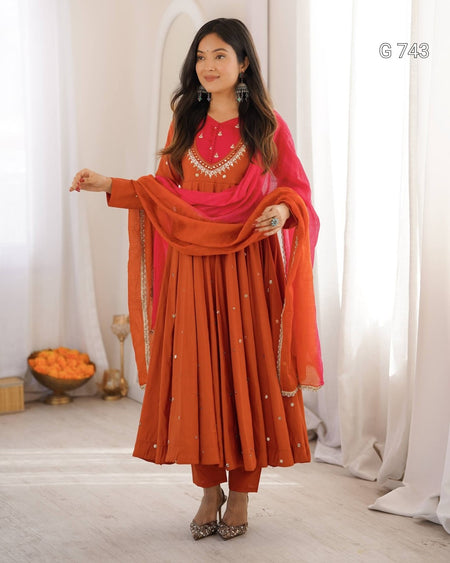 Woman wearing an orange and pink traditional outfit with a red blouse, standing against a white curtain background.