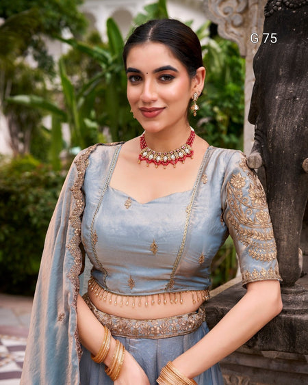 A woman wearing a light blue traditional Indian outfit with gold embroidery, a matching dupatta, and gold jewelry, standing outdoors next to a stone statue.