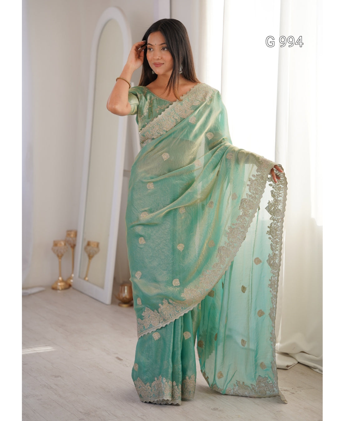 A woman models a mint green saree with gold floral motifs and a silver embroidered border, standing in a brightly lit room with a large mirror and white curtains.