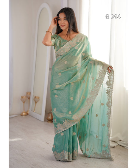 A woman models a mint green saree with gold floral motifs and a silver embroidered border, standing in a brightly lit room with a large mirror and white curtains.