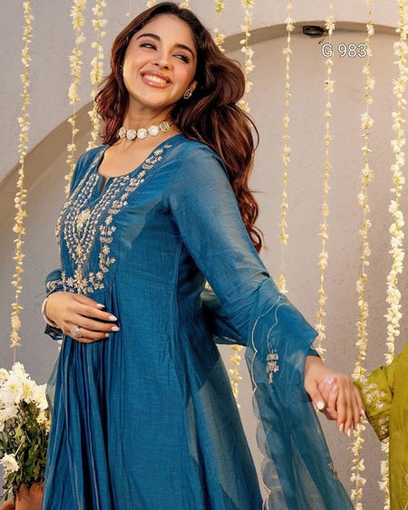 A woman wearing a teal blue traditional Indian salwar kameez outfit with intricate silver embroidery on the chest and sleeves, posed in front of a decorated backdrop with hanging white flowers.
