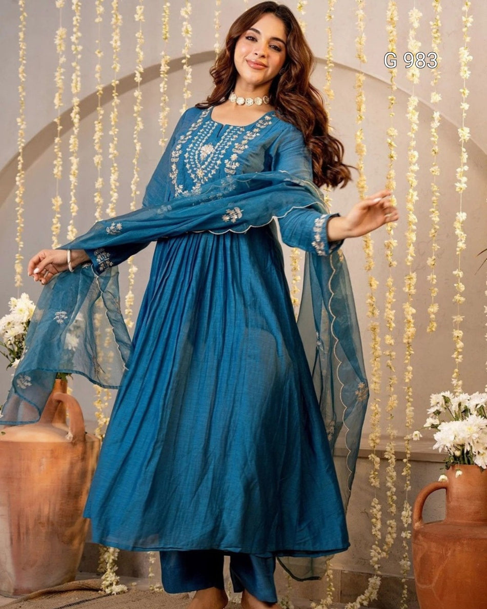 A woman modeling a traditional blue Indian salwar kameez suit with matching dupatta, featuring intricate silver embroidery on the chest and sleeves, set against a decorated backdrop with hanging floral garlands.