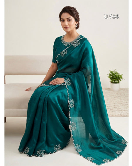 A woman seated on a light beige sofa wearing a teal green saree with white floral embroidery along the border and neckline. The background is a clean indoor setting with a small potted plant on a rug.
