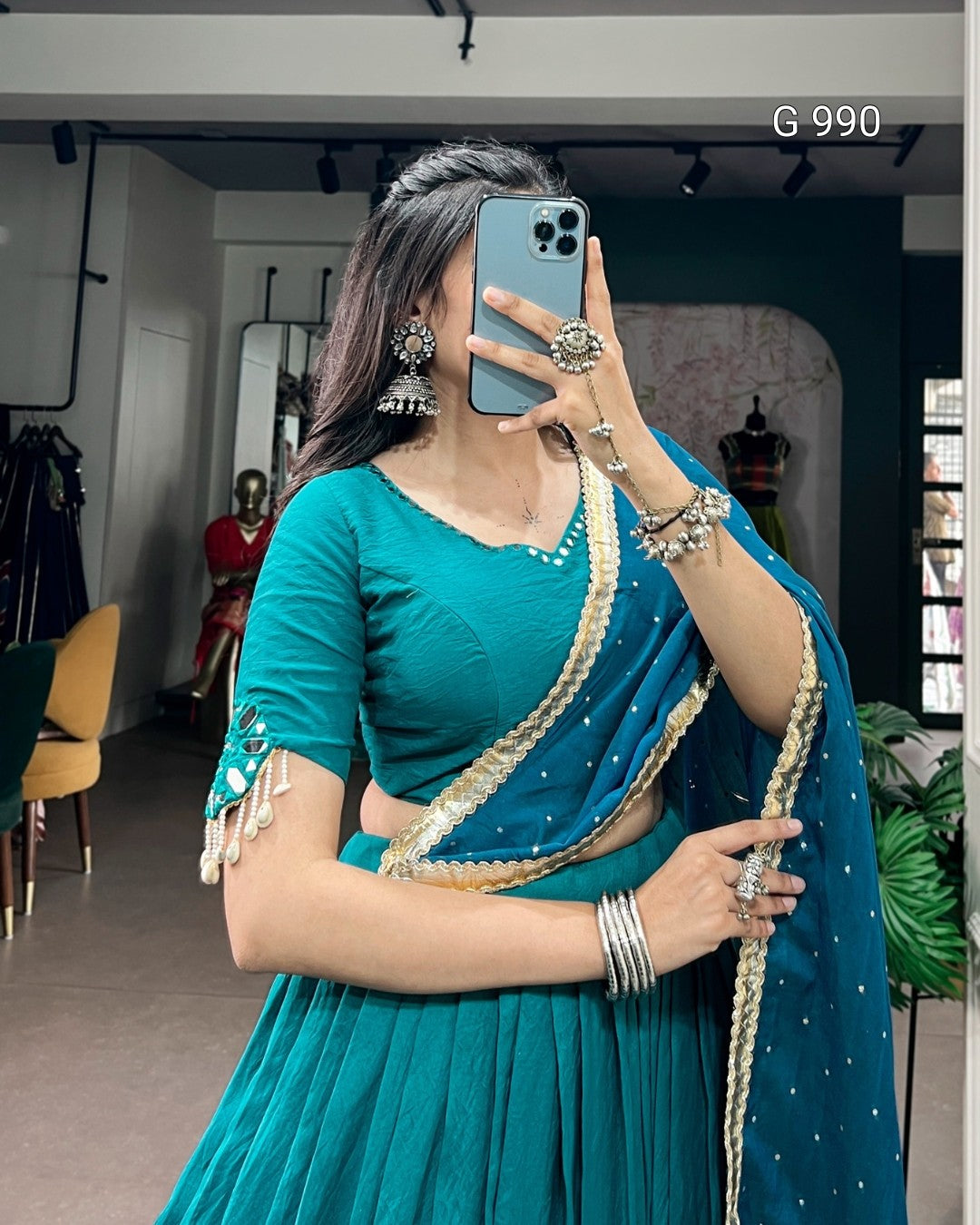 A woman takes a mirror selfie wearing a teal blue traditional outfit with a matching skirt, embellished blouse, and draped dupatta with gold trim. She is holding a blue smartphone in a clothing store setting.